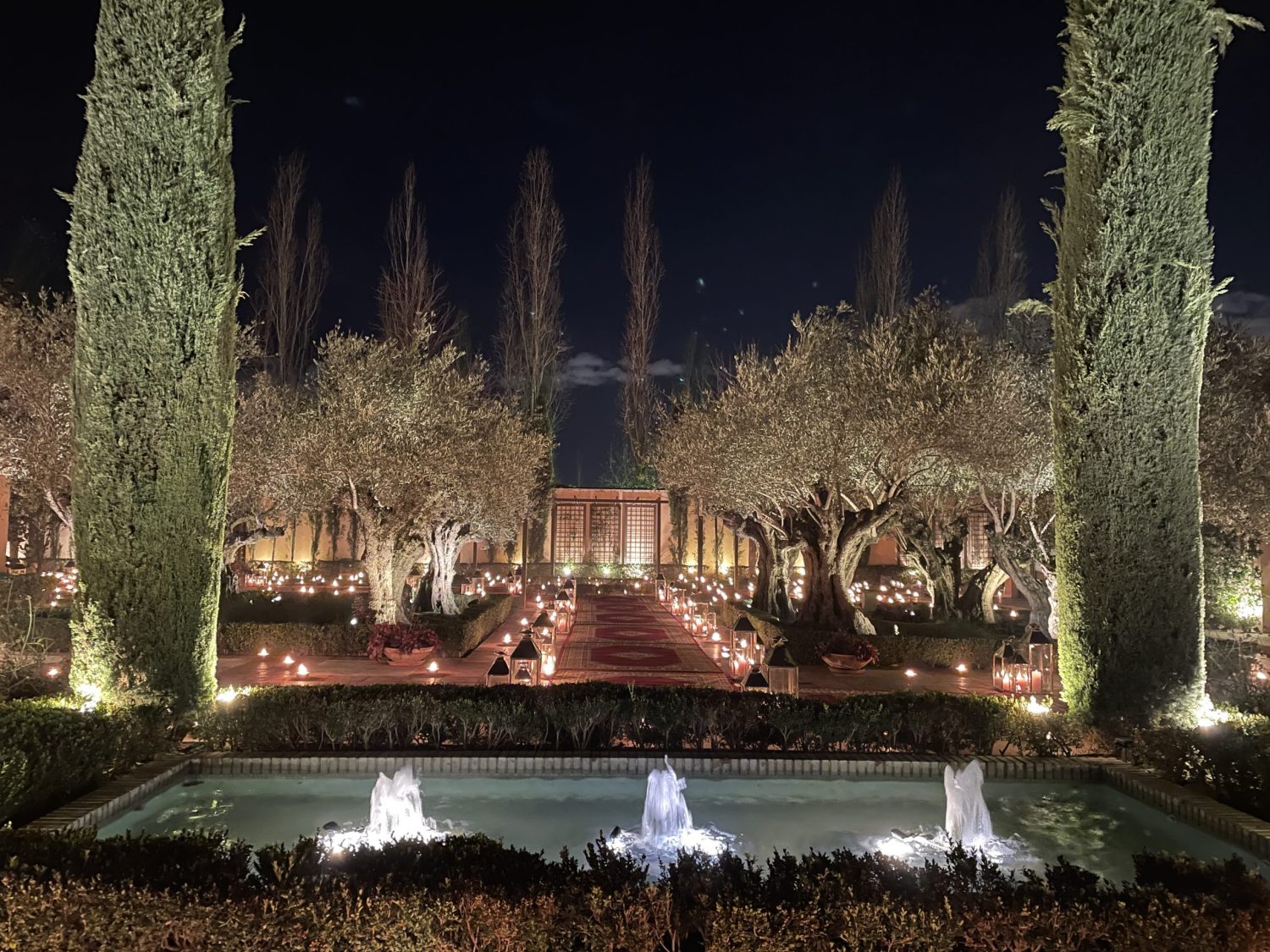 Jardín iluminado de noche con fuentes, olivos y camino decorado con velas y alfombras