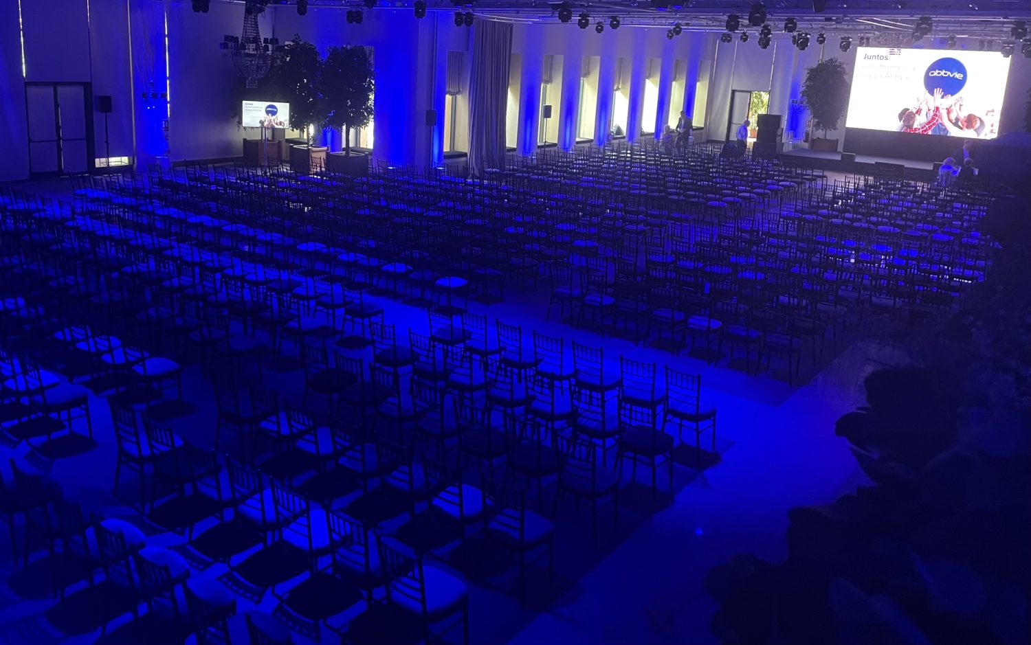 Salón de conferencias con iluminación azul y un gran número de sillas alineadas, preparado para un evento corporativo con un escenario y pantalla en La Quinta de Jarama.
