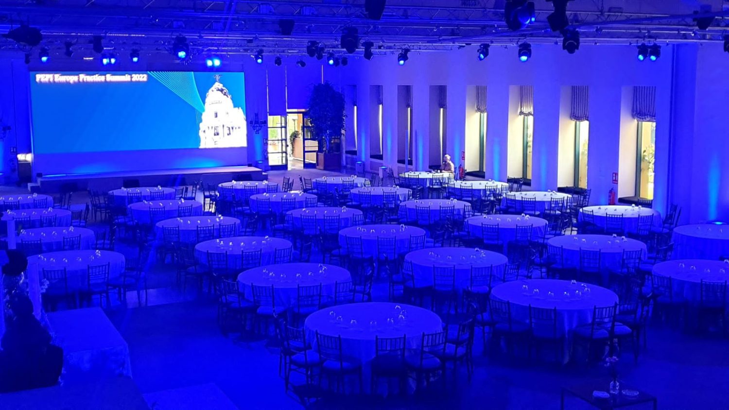 Vista panorámica del salón de eventos en La Quinta de Jarama preparado para la cumbre P2P Europe, con mesas redondas y una gran pantalla iluminada en azul en el escenario.