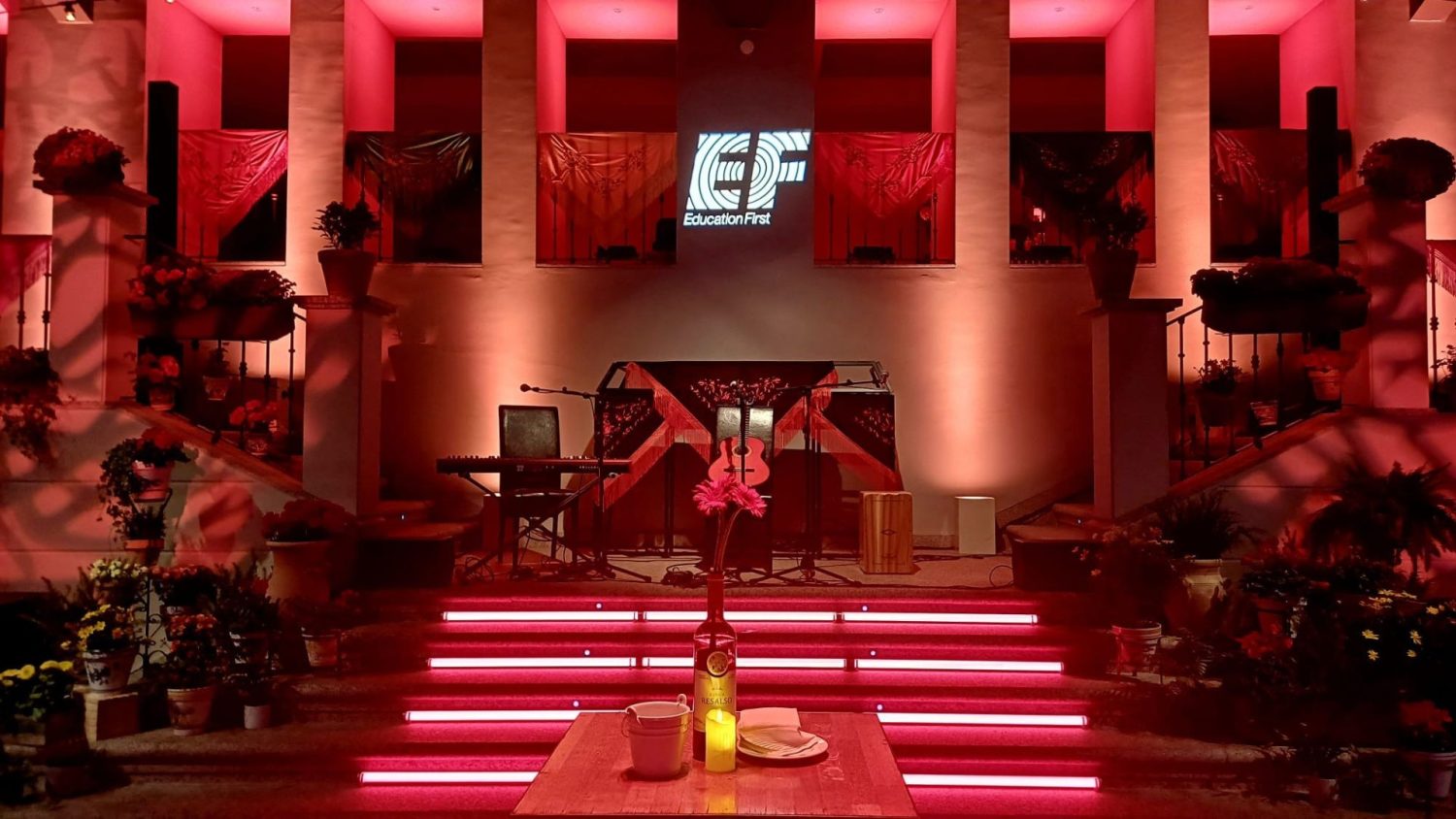 Escenario iluminado en tonos rojos en La Quinta de Jarama para el evento especial con EF. Hay una tarima central preparada para música en directo, instrumentos colocados sobre el escenario y una iluminación ambiental elegante. En primer plano, una mesa decorativa con botella y vela, y al fondo una estructura arquitectónica clásica que crea una atmósfera sofisticada y acogedora
