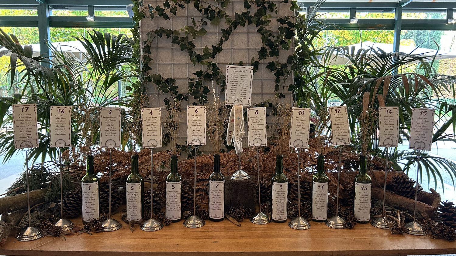 Seating plan decorativo para boda, con tarjetas numeradas sobre soportes metálicos colocadas sobre una mesa rústica, rodeada de vegetación natural, hiedra, piñas y plantas verdes, creando un ambiente elegante y orgánico para la asignación de mesas