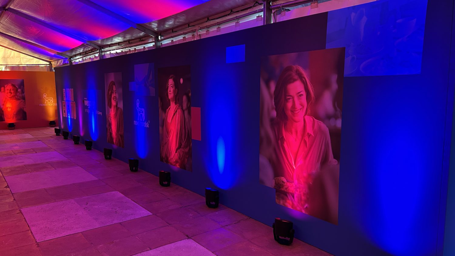Pasillo con paneles informativos y retratos iluminados en azul en el evento Novo Nordisk en la Quinta de Jarama