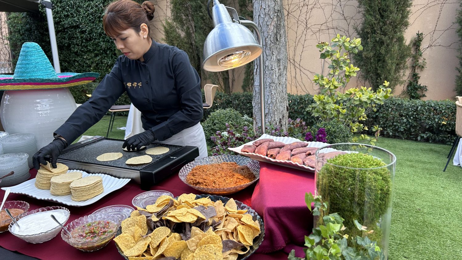Cocinera uniformada colocando pequeñas tortillas de maíz en una plancha caliente de buffet en el jardín, con una gran variedad de ingredientes mexicanos listos para servir.