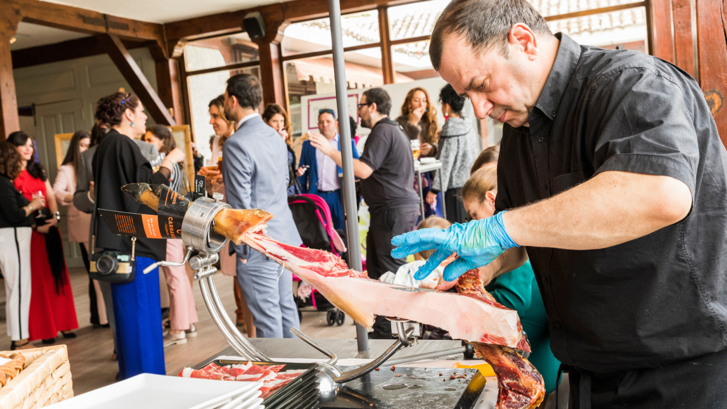 Un hombre con camisa negra y guantes azules cortando una pata de jamón en un soporte metálico, con invitados de boda vestidos de fiesta conversando en el fondo.