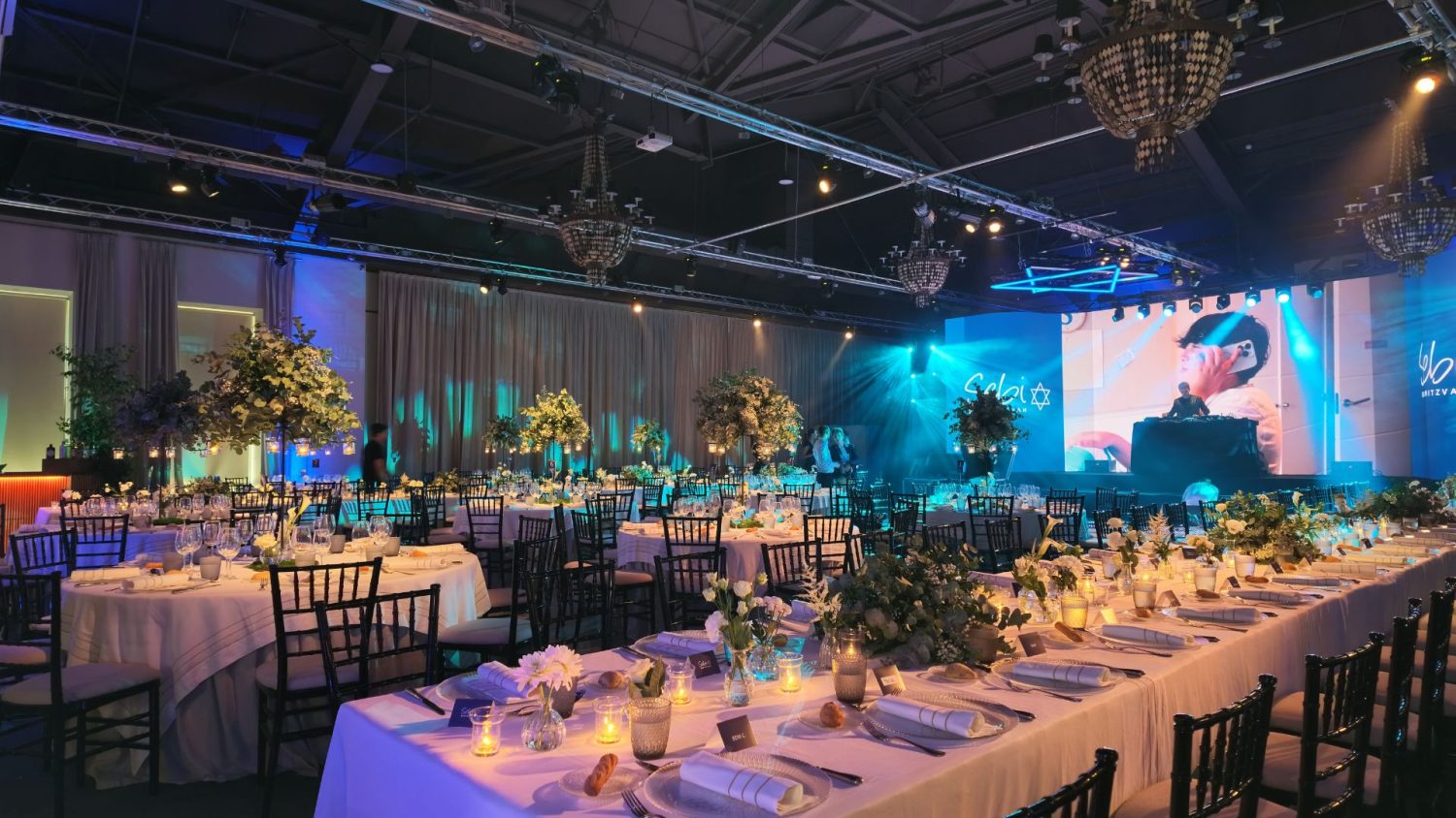 Elegante montaje de banquete para celebración de Bar Mitzvá en La Quinta de Jarama, Madrid. El salón destaca por su cuidada iluminación en tonos azules, mesas largas y redondas con decoración floral, y un escenario con pantalla gigante animando la fiesta.