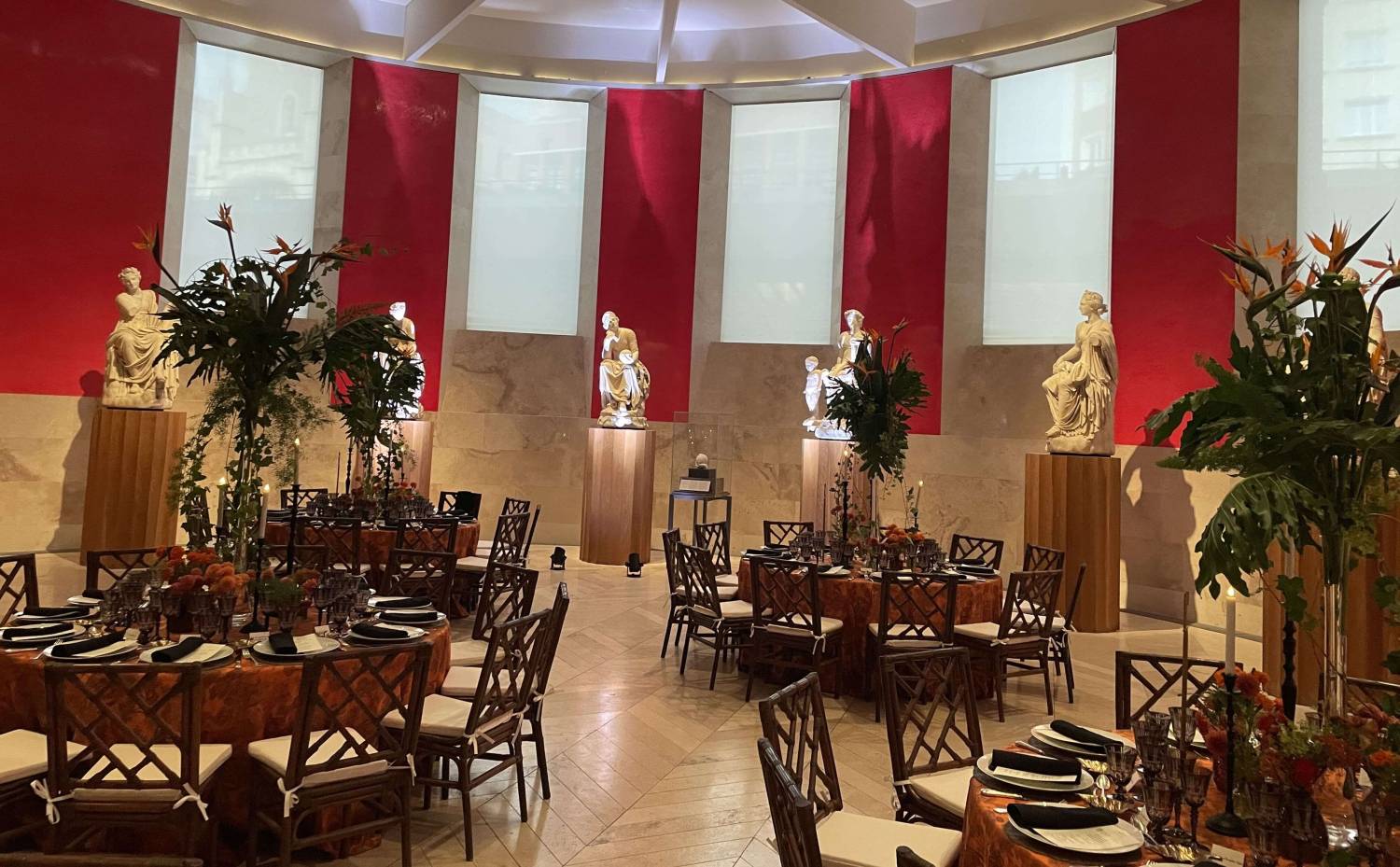 Mesas redondas con mantelería naranja y centros vegetales altos, rodeadas de esculturas en el Museo Nacional del Prado.